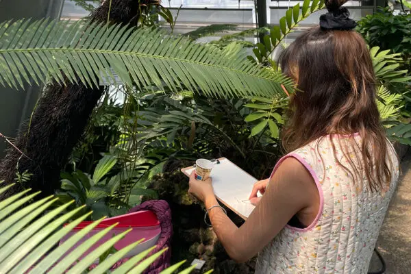 A person standing in a greenhouse. They are holding a paper block on which they are drawing the plants they are seeing.