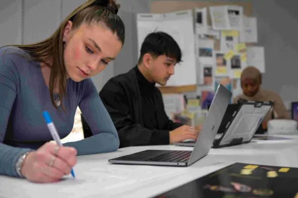 Students using laptops and writing in class, with a notice board in the background