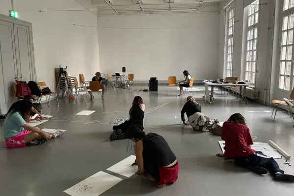 8 people sitting on the floor of a big room, spread out. Each of them has one or more pieces of paper in front of them, on which they are drawing.