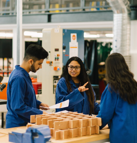 Three students in discussion, situated in a BCU engineering workshop, all wearing blue lab coats