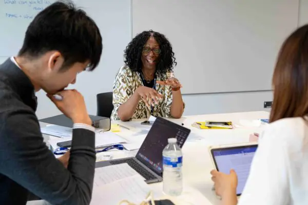 Lecturer smiles mid discussion with two students, both whom have laptops on a desk